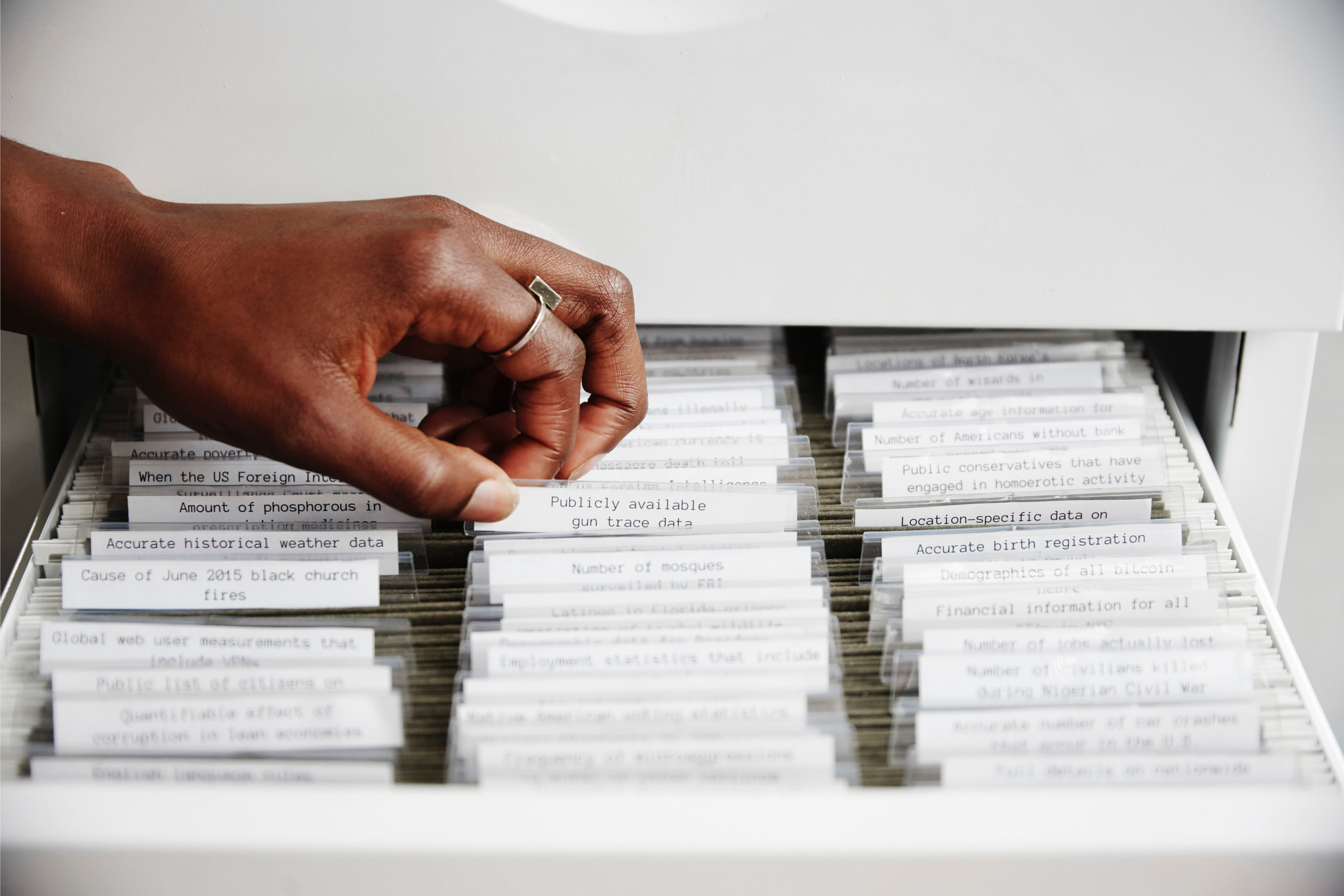 A feminine hand with darker skin reaches into a filing cabinet with many files inside, each with tabbed labels. The hand is reaching for “Publicaly available gun trace data”. The image is from MIMI ỌNỤỌHA’s website and is of ‘The Library of Missing Datasets’ (2016)